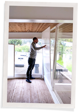 A person is installing a large window insert while wearing a grey sweater and dark pants.