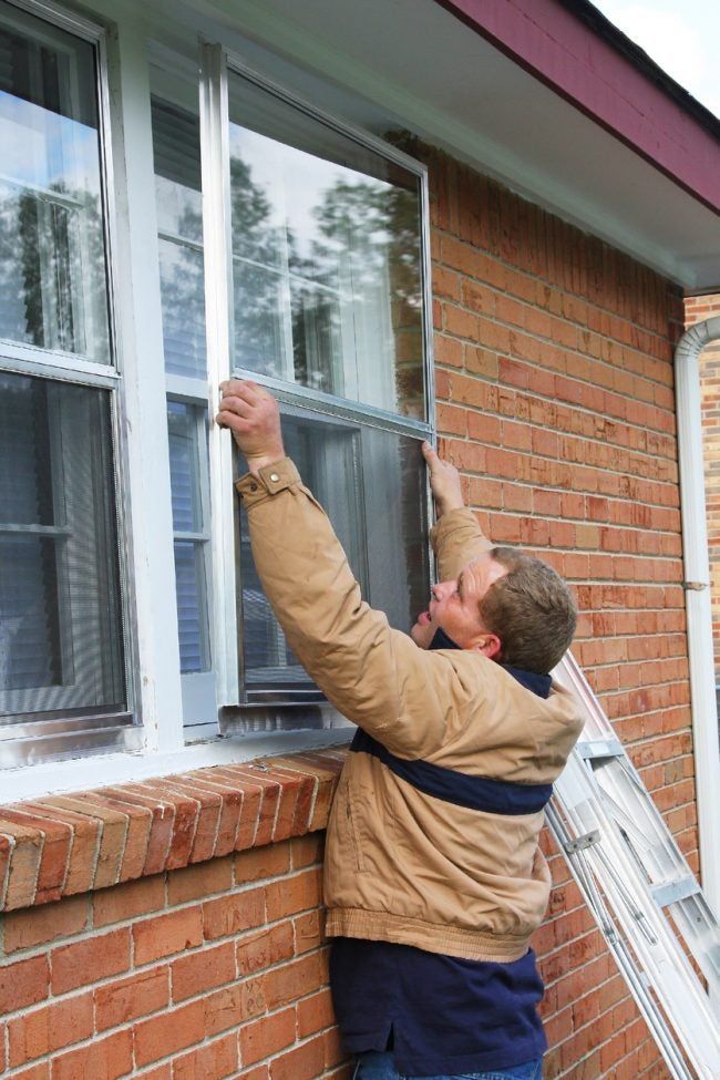 A person wearing a tan coat is installing storm window on a brick home.