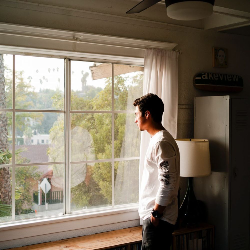A person is standing inside of a house, staring out a large white window, looking out onto the neighborhood street.