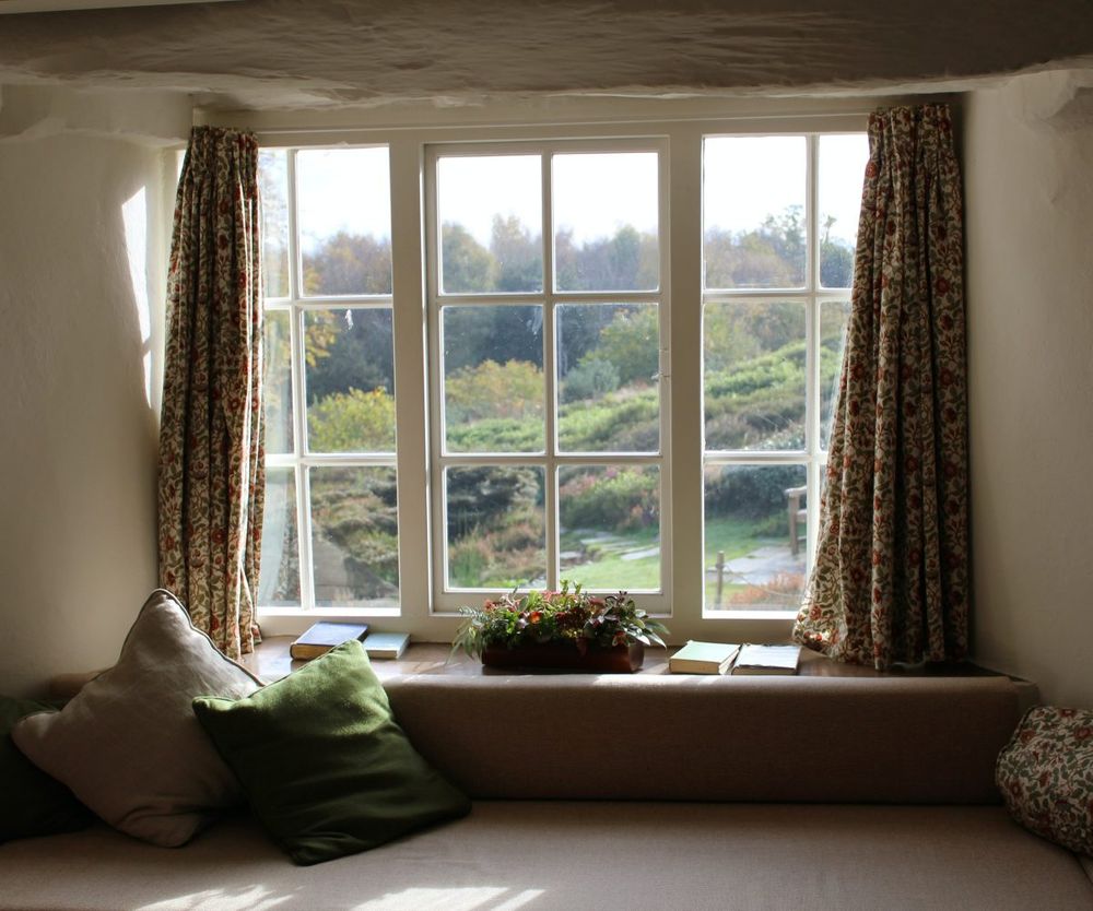 A window with a sitting area overlooks a beautiful outdoor landscape with various trees and flowers.