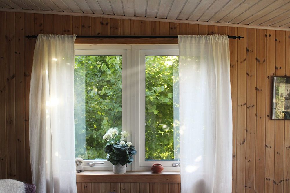 Window looking out into trees. There are white curtains on both sides of the window. The window sill has an orange bowl and a flower vase on it.