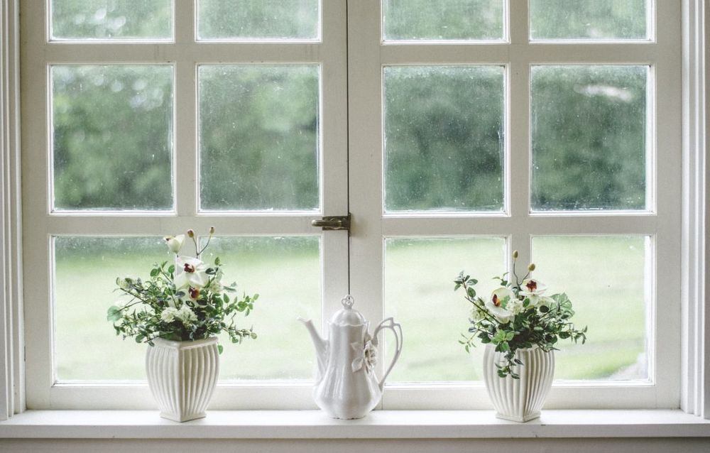 Two windows have white painted panes with a window lock on the bottom half of the window. There are two plants sitting on the window sill and a teapot in between them.