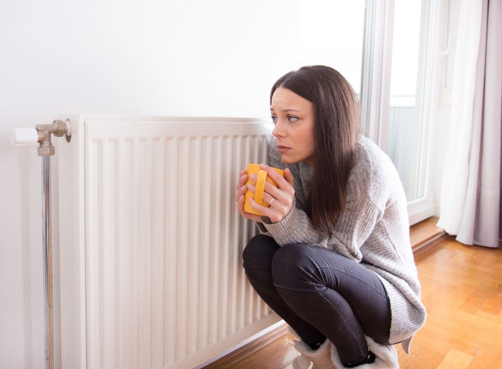 A person is crouched next to a heater, holding an orange coffee mug.