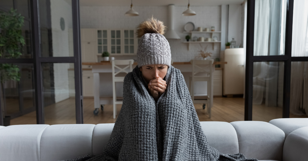 A woman sitting in a blanket and a woolen hat in her living room,
			warming up her hands.