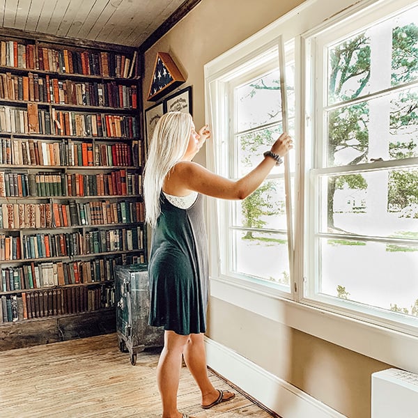 woman installing Indow window inserts to reduce noise next to bookshelves