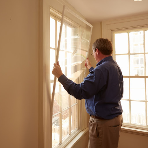 A person wearing a blue shirt and khakis installs a window insert in a window frame.